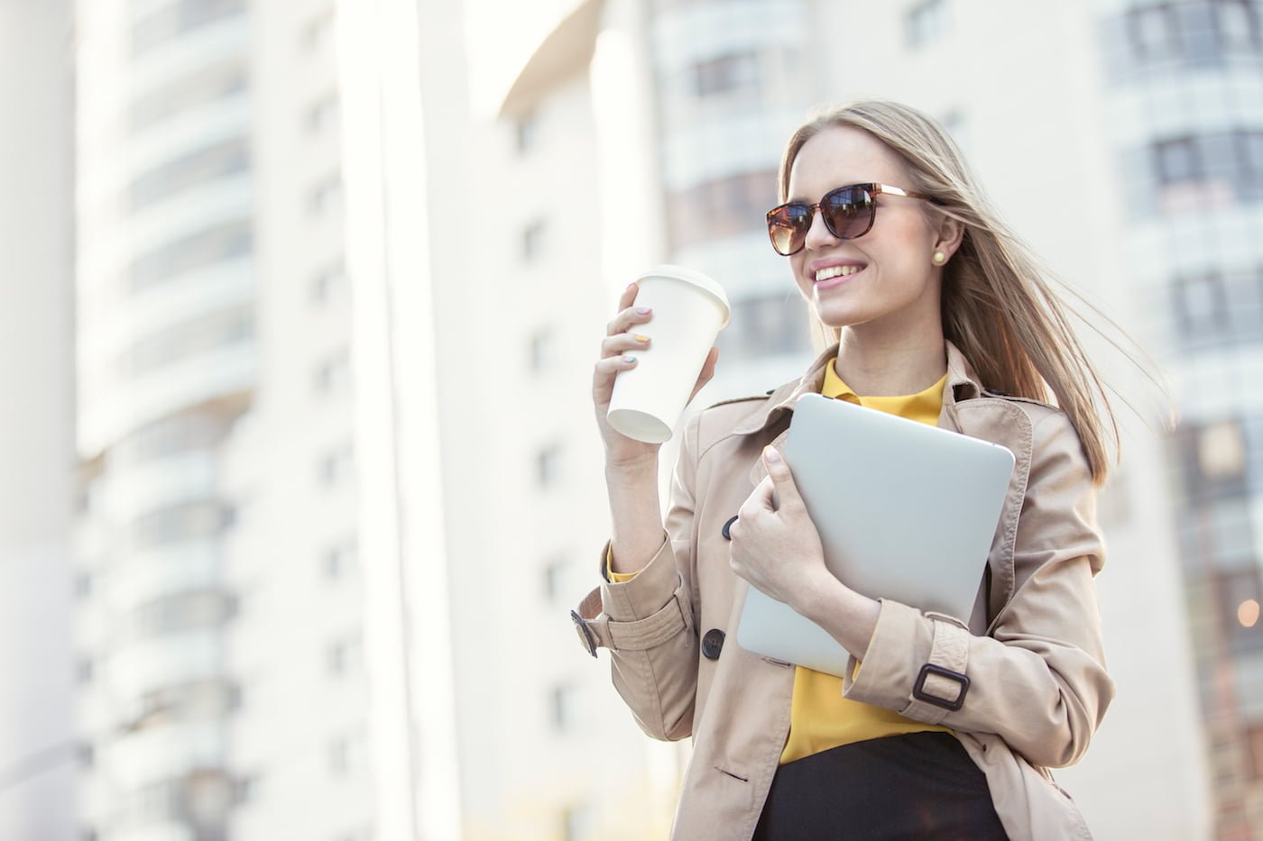 Beautiful young businesswoman with a disposable coffee cup, drinking coffee, and holding tablet in her hands against urban city background.